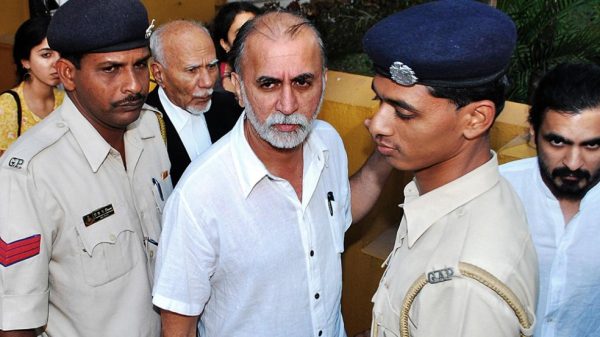 Bearded man in a white shirt escorted by two uniformed police officers while onlookers observe nearby.