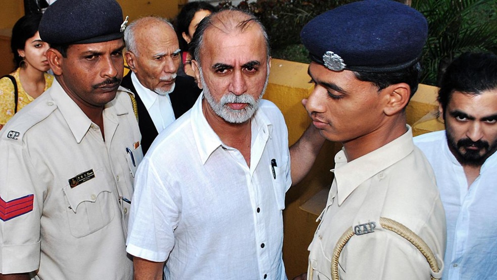 Bearded man in a white shirt escorted by two uniformed police officers while onlookers observe nearby.