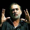 Male speaker with gray beard on a dark stage, hands raised in a gesturing pose while talking.