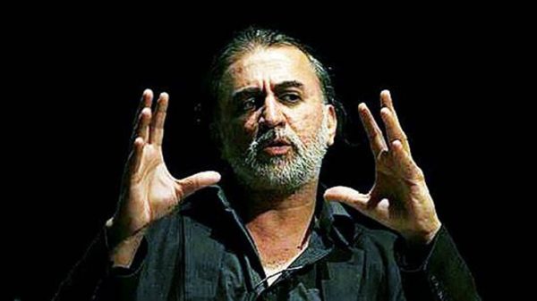 Male speaker with gray beard on a dark stage, hands raised in a gesturing pose while talking.
