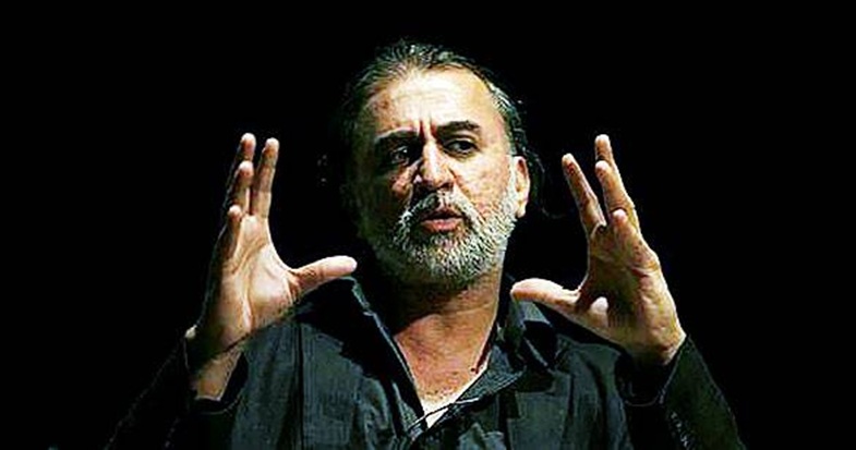 Male speaker with gray beard on a dark stage, hands raised in a gesturing pose while talking.