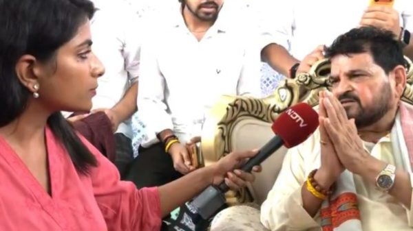 Female reporter in a pink top interviews a bearded man in traditional attire; hands folded in prayer as he speaks.