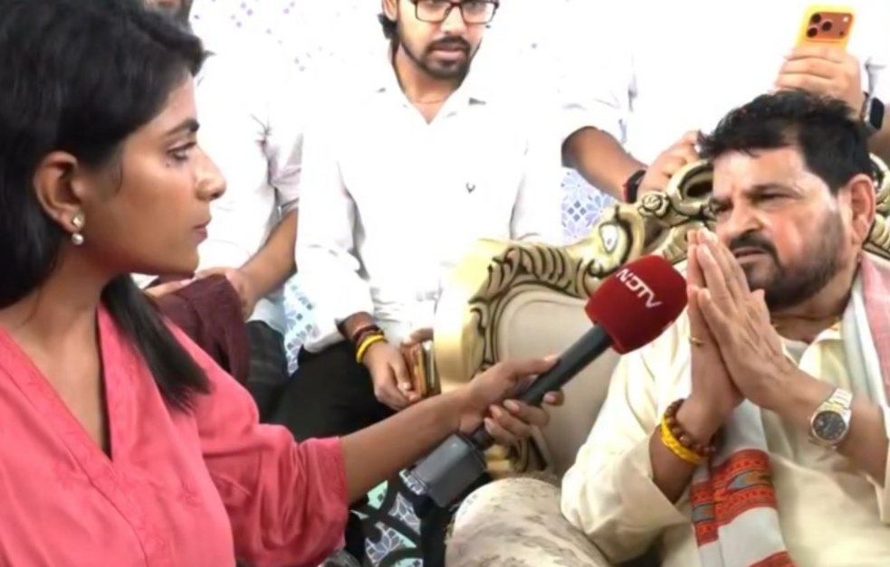 Female reporter in a pink top interviews a bearded man in traditional attire; hands folded in prayer as he speaks.
