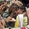 Young boy surrounded by soldiers, distressed as a few hands hold him by the head amid a tense scene