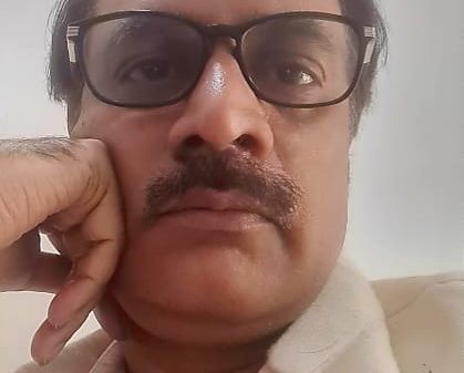 Man with rectangular glasses and mustache resting his chin on his hand, looking at the camera in a close-up shot.