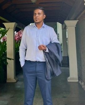 Man in a light blue dress shirt and navy trousers stands on a covered walkway, holding a dark suit jacket.