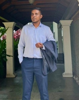 Man in a light blue dress shirt and navy trousers stands on a covered walkway, holding a dark suit jacket.