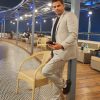 Man in a light gray suit leaning on a wicker chair at an outdoor restaurant at night, holding a phone.