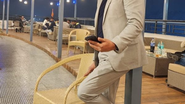 Man in a light gray suit leaning on a wicker chair at an outdoor restaurant at night, holding a phone.