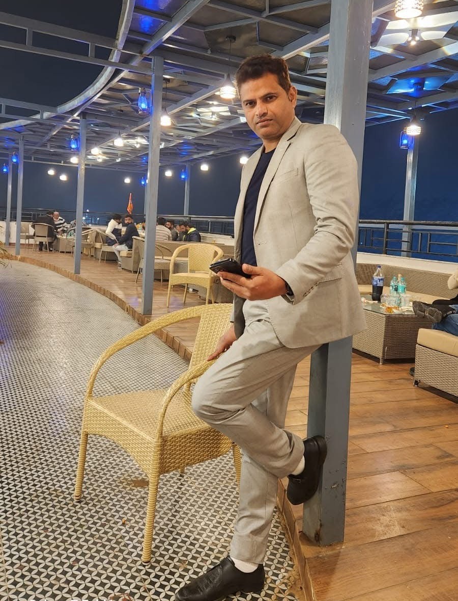 Man in a light gray suit leaning on a wicker chair at an outdoor restaurant at night, holding a phone.