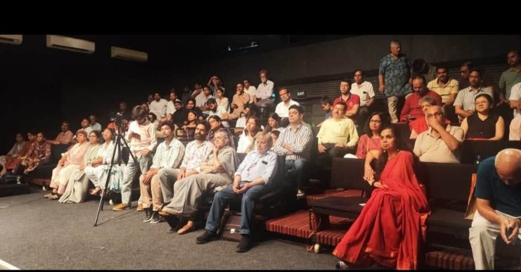 viren ji_1 | No. 1 Indian Media News Portal Audience seated in a dark auditorium watching a talk; in the front row a woman in a red sari speaks into a microphone, others sit nearby, with a tripod-mounted camera visible at the left.