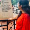 Woman in a red outfit standing at a railing, reading a tall stone plaque with Hindi text outdoors.