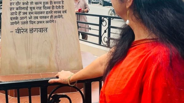 Woman in a red outfit standing at a railing, reading a tall stone plaque with Hindi text outdoors.