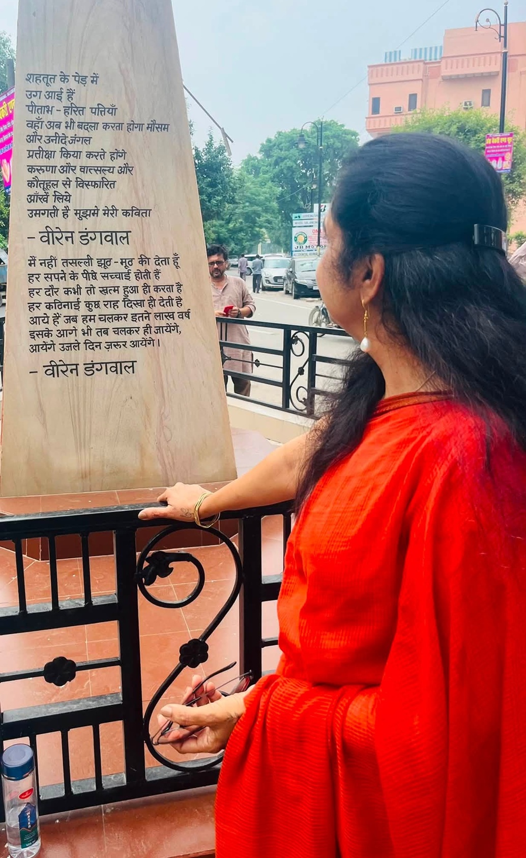 Woman in a red outfit standing at a railing, reading a tall stone plaque with Hindi text outdoors.