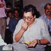 Man in a light gray shirt and glasses sits at a table, biting into a chip while holding more chips; several people in casual attire smile in the background at a social gathering.