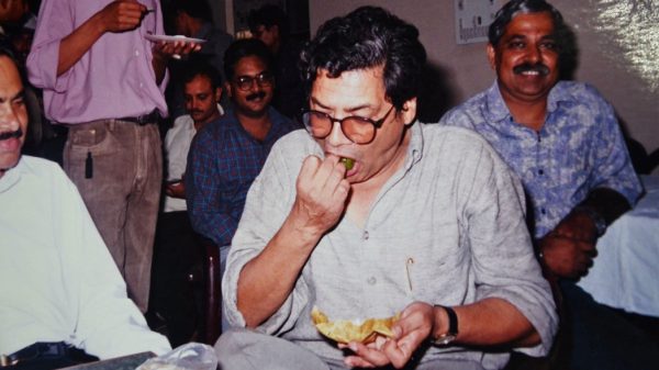 Man in a light gray shirt and glasses sits at a table, biting into a chip while holding more chips; several people in casual attire smile in the background at a social gathering.