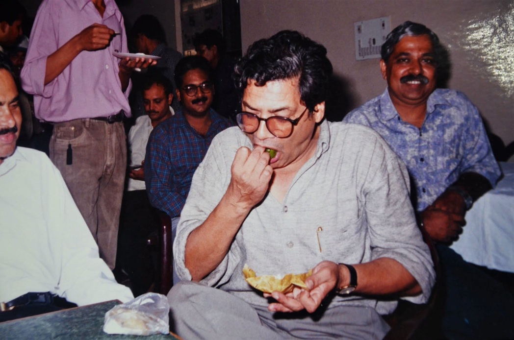 Man in a light gray shirt and glasses sits at a table, biting into a chip while holding more chips; several people in casual attire smile in the background at a social gathering.