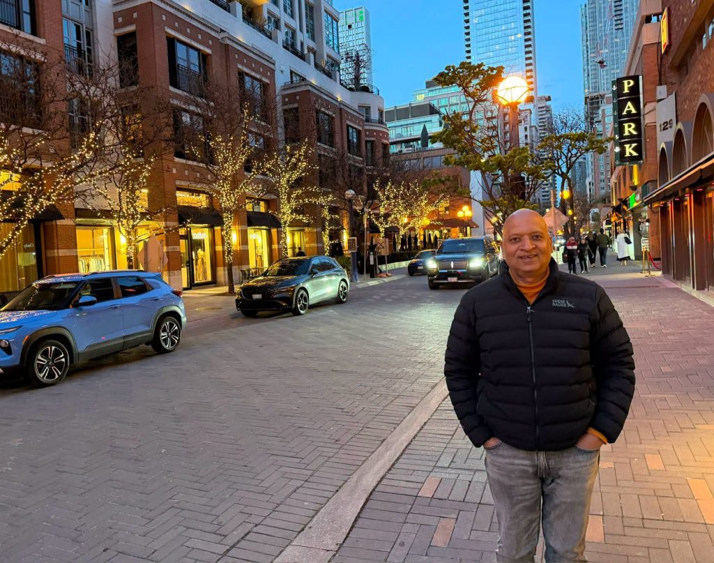Smiling man in a black puffer jacket stands on a brick-paved city street at dusk, storefronts and string-lit trees behind him.