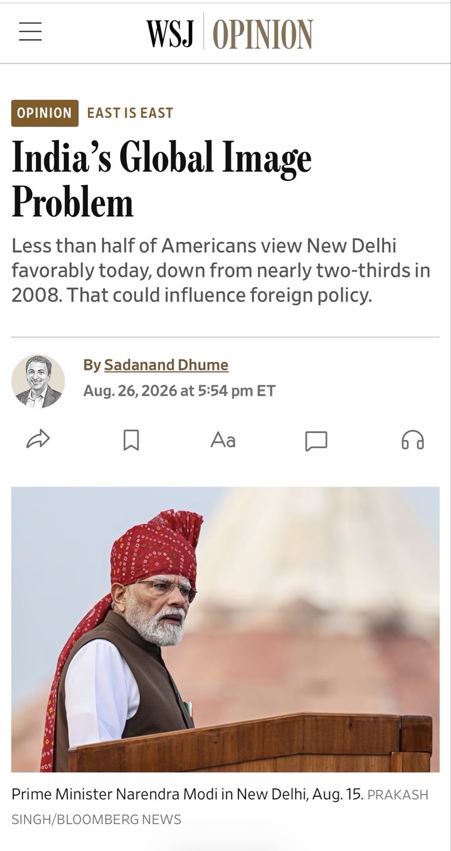 Photograph of a bearded man in a red headscarf speaking at a wooden podium during a public address in New Delhi, with a blurred backdrop behind him.