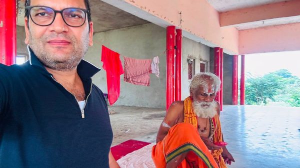 Man in the foreground wearing glasses takes a selfie; in the background a bearded monk in orange robes sits on a mat under pink columns.