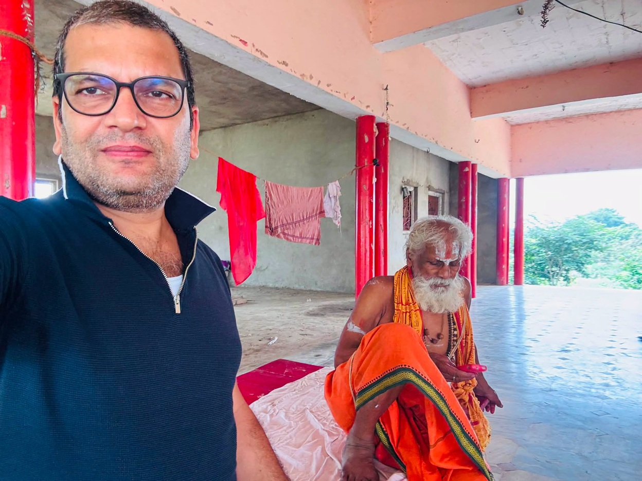 Man in the foreground wearing glasses takes a selfie; in the background a bearded monk in orange robes sits on a mat under pink columns.