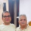 Two men posing for a selfie indoors; the man on the left wears glasses and a white shirt, the man on the right has a gray mustache and a light patterned shirt.
