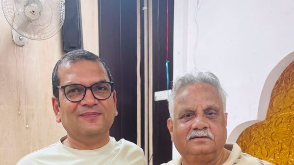 Two men posing for a selfie indoors; the man on the left wears glasses and a white shirt, the man on the right has a gray mustache and a light patterned shirt.