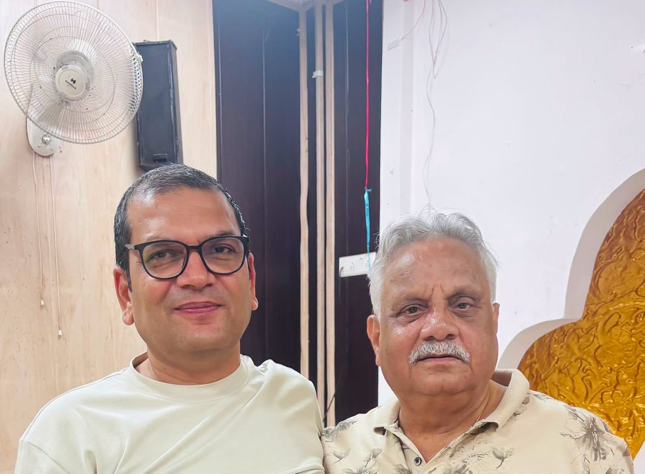 Two men posing for a selfie indoors; the man on the left wears glasses and a white shirt, the man on the right has a gray mustache and a light patterned shirt.