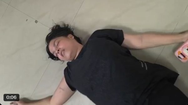 Woman in a black shirt lying on a light tile floor, eyes closed, arms spread, holding a pink object in her right hand.