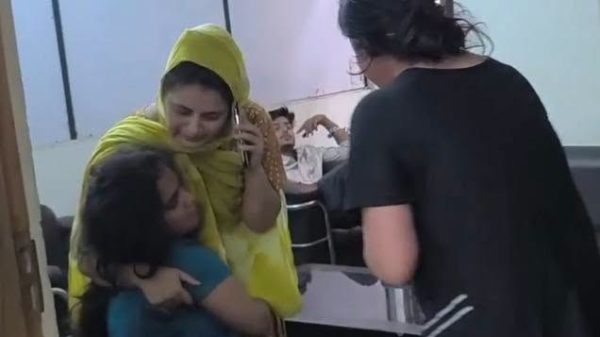 Woman in a yellow hijab comforts a crying child who is being hugged by another woman in a clinic waiting room as a staff member approaches.