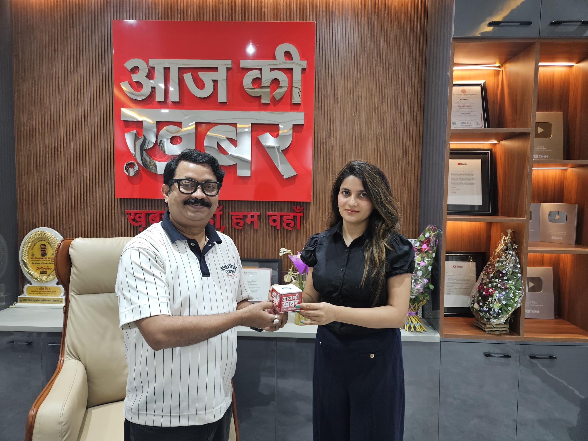 Two people exchange a red mug in a showroom, with a wooden wall and award shelf in the background.