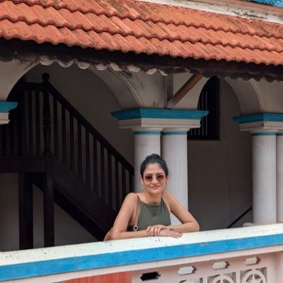 7c5bf8ce-09d8-4b02-af76-fb0b51caacf2 | No. 1 Indian Media News Portal Smiling woman in a green sleeveless top leaning on a blue-and-white railing of a courtyard balcony, with arches and a staircase in the background.