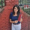Smiling woman in a blue top and white pants leaning against a red brick wall, holding a red hardcover book.