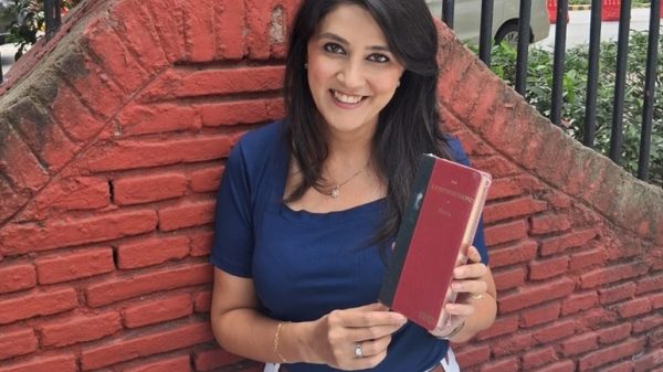 Smiling woman in a blue top and white pants leaning against a red brick wall, holding a red hardcover book.
