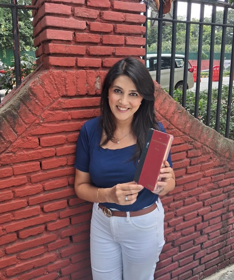 Smiling woman in a blue top and white pants leaning against a red brick wall, holding a red hardcover book.
