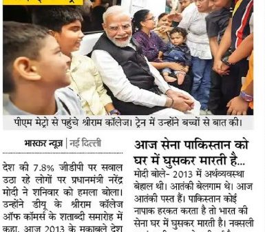 Prime Minister Modi sits with children and adults at a campus event, shaking hands and talking with them in a crowded setting in India.