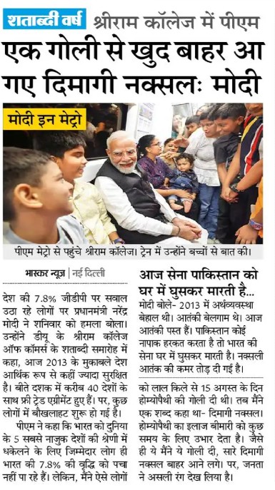 Prime Minister Modi sits with children and adults at a campus event, shaking hands and talking with them in a crowded setting in India.
