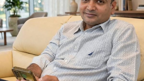 Man relaxing on a beige sofa, holding a smartphone and looking at the camera in a bright living room.