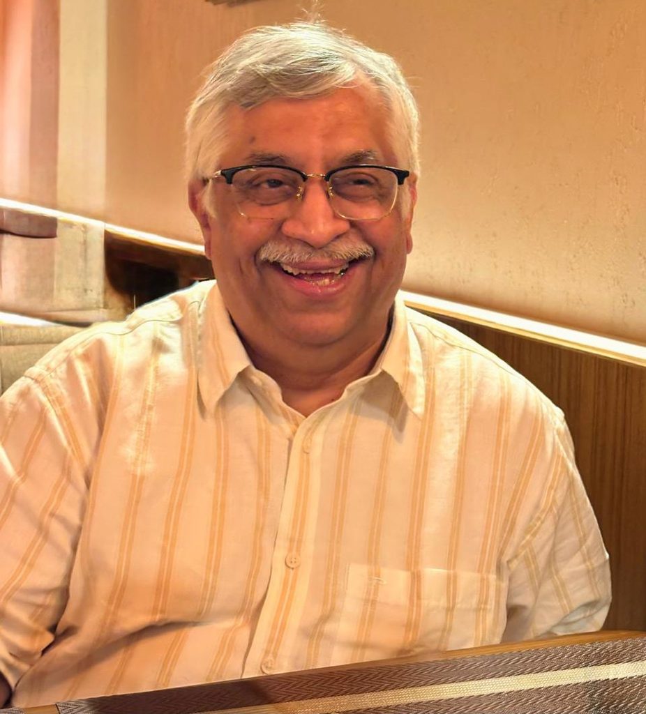Smiling older man with gray hair and glasses wearing a light striped shirt, seated in a booth at a restaurant.