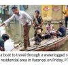 Residents move through a flooded street in Varanasi by boat, with several people aboard and others on the waterlogged bank.