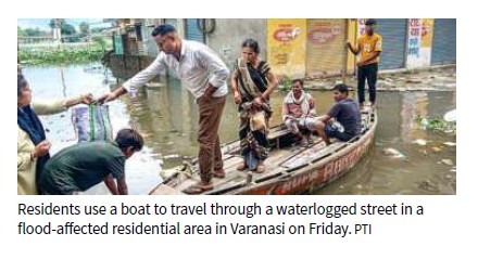 Residents move through a flooded street in Varanasi by boat, with several people aboard and others on the waterlogged bank.