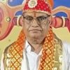 Man in traditional ceremonial attire with a red jeweled turban and orange scarf, standing before a colorful backdrop.