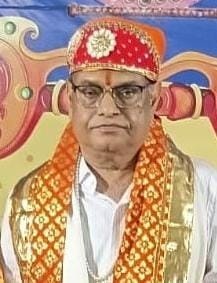 Man in traditional ceremonial attire with a red jeweled turban and orange scarf, standing before a colorful backdrop.