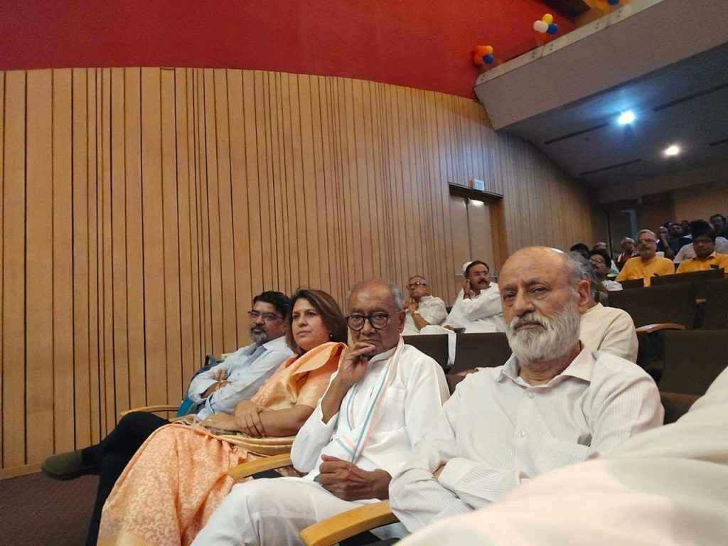 akhlaq_1 | No. 1 Indian Media News Portal Audience seated in a conference hall with wood-paneled walls; foreground shows an elderly man in a white shirt, beard, and others beside him. Free-form group of attendees in the background, some in traditional clothing.