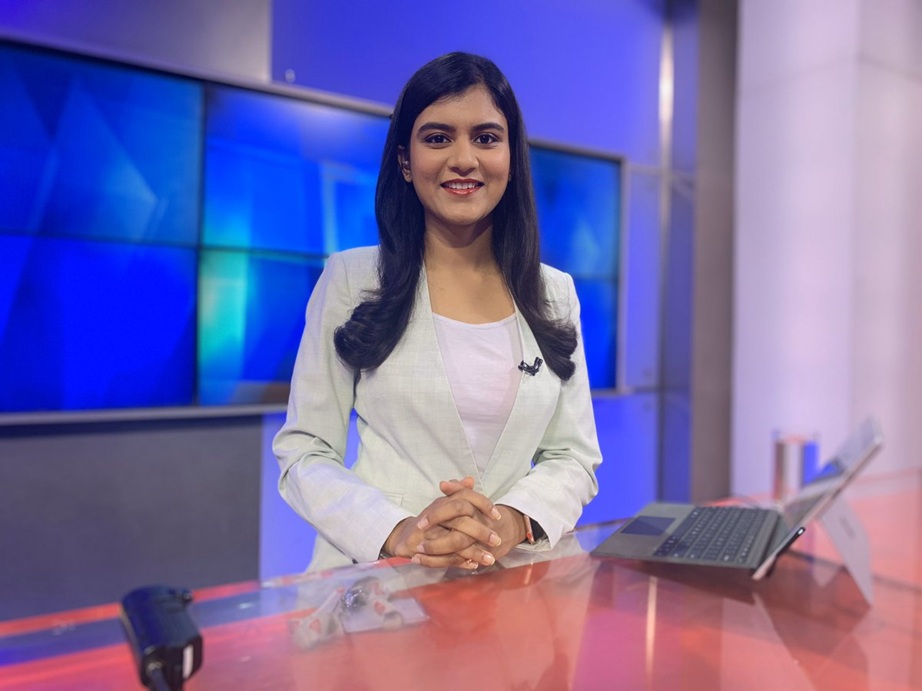 akshita | No. 1 Indian Media News Portal Female news anchor in a white blazer sits at a glass desk with a laptop, smiling at the camera in a studio set with blue panels behind her.