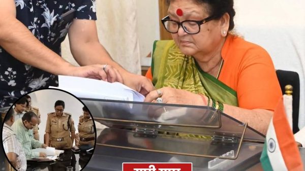 Woman in orange saree and glasses signs documents at a desk while a man assists; Indian flag in foreground and a round news logo in the corner; inset shows officials reviewing papers with police present.