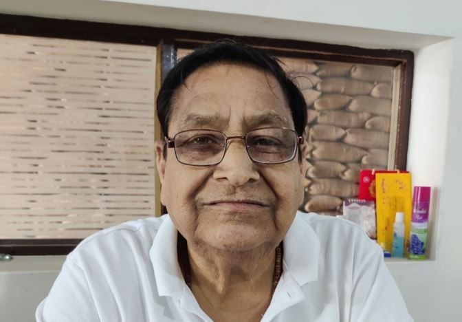 amritlal ji | No. 1 Indian Media News Portal Close-up portrait of an elderly man with glasses wearing a white polo shirt, looking at the camera with a gentle smile, indoors with shelves and items in the background.