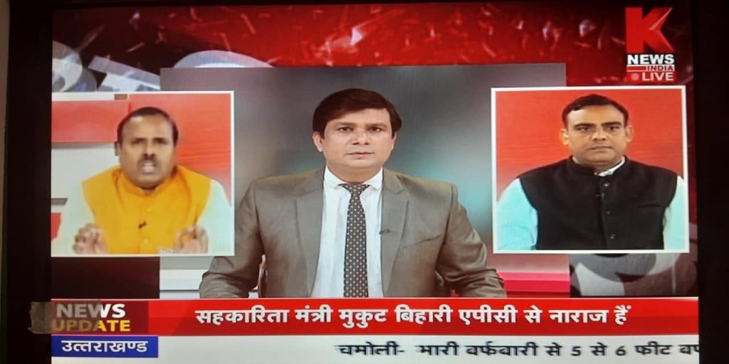 anchor om | No. 1 Indian Media News Portal News anchor in a gray suit reports live, flanked by two inset guest panels on a red-badged studio background, with a Hindi ticker at the bottom and K News India Live logo top-right.