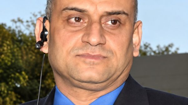 Close-up of a bald man in a blue shirt and dark suit wearing a headset with a microphone outdoors.