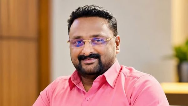Smiling man with short black hair, beard, and glasses wearing a pink button-down shirt indoors.